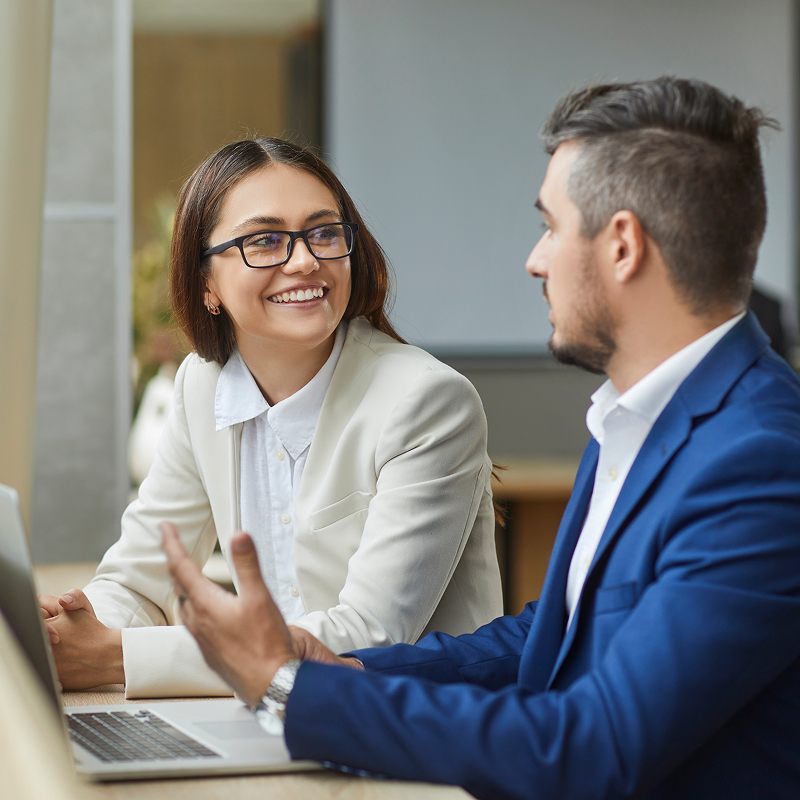 shutterstock_2486613543-img Two young smiling company employees man and woman sitting with laptop having discussion talking